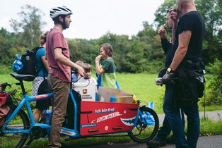 Freight bike in the green. Surrounded by children and adults eating ice cream. Crate of Lammsbräu and moving box in the cargo bike. 