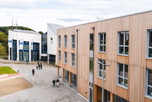 Im Hintergrund ist das blau-weiße Hauptcampusgebäude zu sehen, im Vordergrund der Holzbau der UW/H.