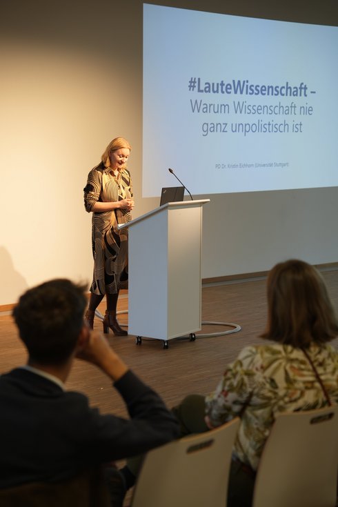 A woman stands at a desk and speaks into a microphone.