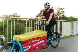 Lachender Mann fährt mit Helm Lastenrad über eine Brücke. 