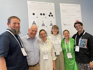 Six people stand in front of science posters and smile at the camera.