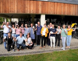 Eine Gruppe von ca. 25 Personen steht vor einem Holzgebäude und jubelt.