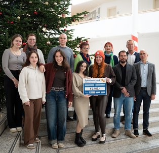 Eine Menschengruppe steht auf einer Treppe vor einem Weihnachtsbaum. Ein großer symbolischer Scheck wird von zwei Personen gehalten.