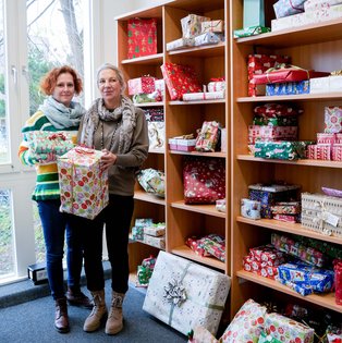 Team der Info-/Poststelle mit den Geschenken (Foto: UW/H) Zwei Frauen stehen vor einem Regal voller verpackter Weihnachtsgeschenke.