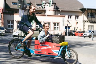 Laughing man riding cargo bike with laughing woman inside and suitcase/luggage in front of a building that could be a train station. 