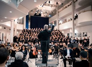 Foto eines Dirigenten der mit dem Rücken zum Publikum steht. Hinter ihm sitzt das Orchester und der Chor.