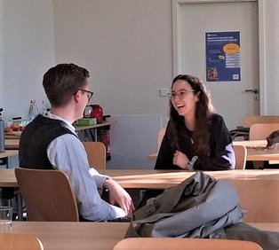Ein Mann und eine Frau sitzen sich in einem Seminarraum im Gespräch gegenüber, die Faru lacht
