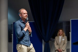 Jonas Redecker (Photo: UW/H | Michael Schwettmann) A man speaks into a microphone, a woman can be seen blurred in the background.
