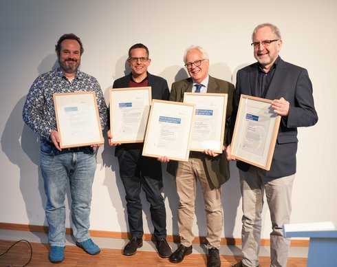 Four men smile at the camera and hold certificates in the picture.