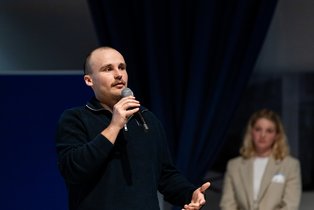 Severin Pfeil (Photo: UW/H | Michael Schwettmann) A man speaks into a microphone, a woman can be seen blurred in the background.